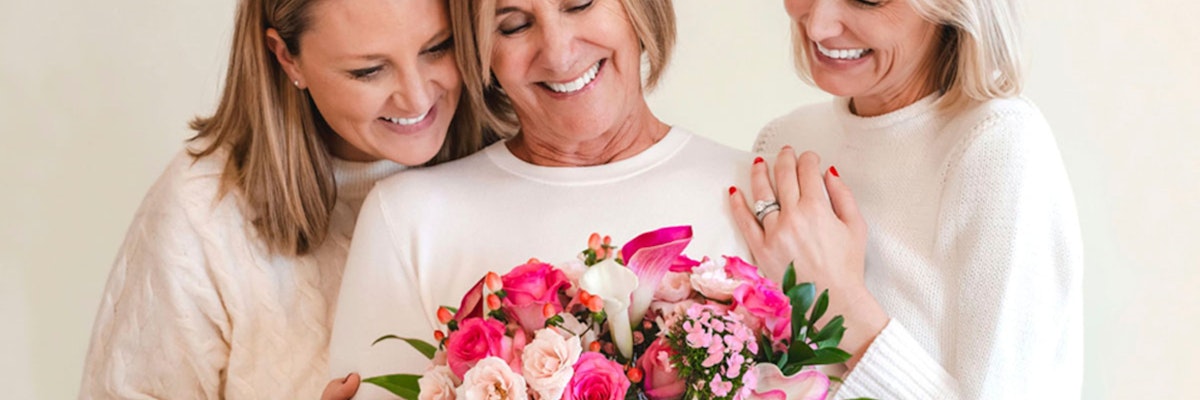 Three women joyfully celebrate together, holding a vibrant bouquet of pink flowers.