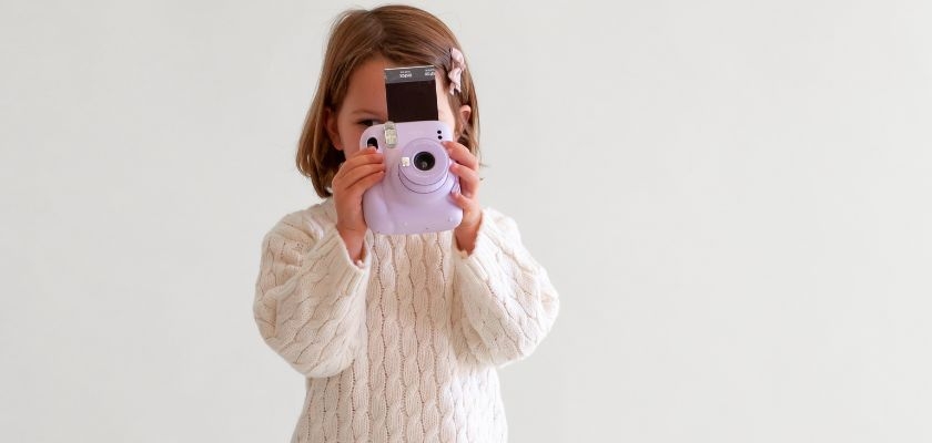 A smiling child holding a cute purple instant camera against a plain background.