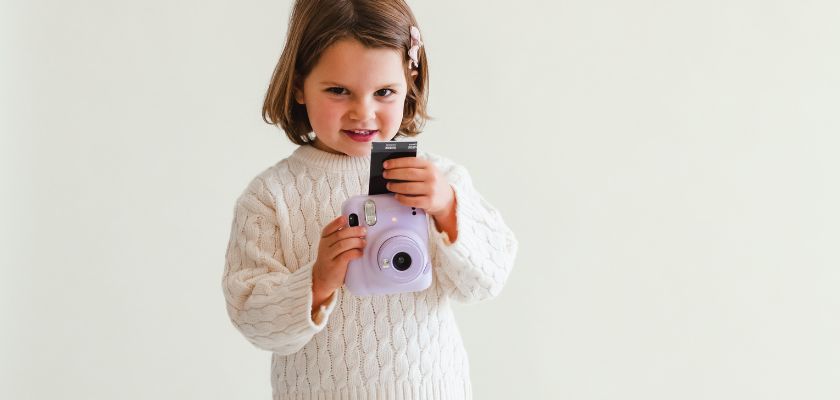 A cheerful girl in a cozy sweater holding a lavender camera, ready to capture moments.