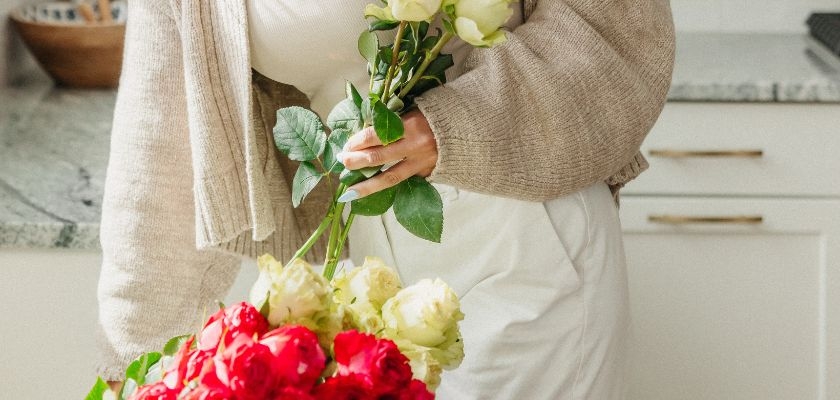 A woman in a cozy outfit holds a vibrant bouquet of red and cream roses in her kitchen.