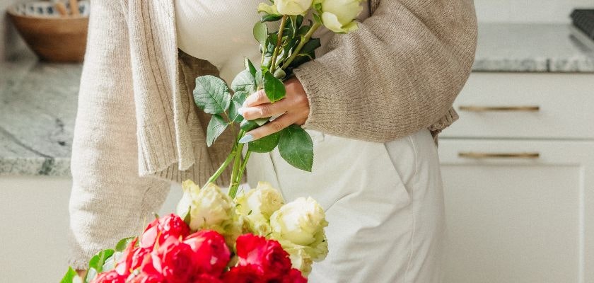 A woman in a cozy outfit holds a vibrant bouquet of red and cream roses in her kitchen.