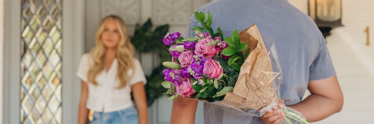 A person holding a vibrant bouquet of pink and purple flowers, greeting someone at the door.