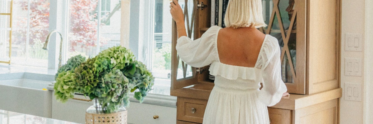 A woman in a white dress stands by a cabinet, admiring a lush green floral arrangement.