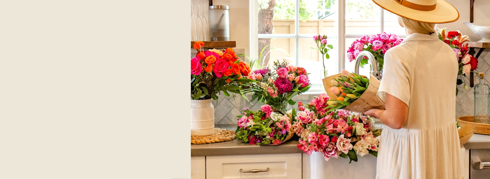 A woman wearing a hat arranges vibrant flower bouquets in a bright kitchen setting.
