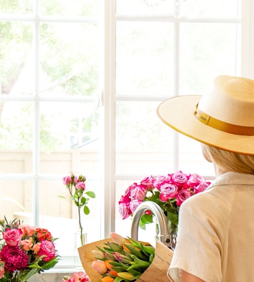 A woman in a sun hat admires vibrant flowers by a bright window, creating a cheerful ambiance.
