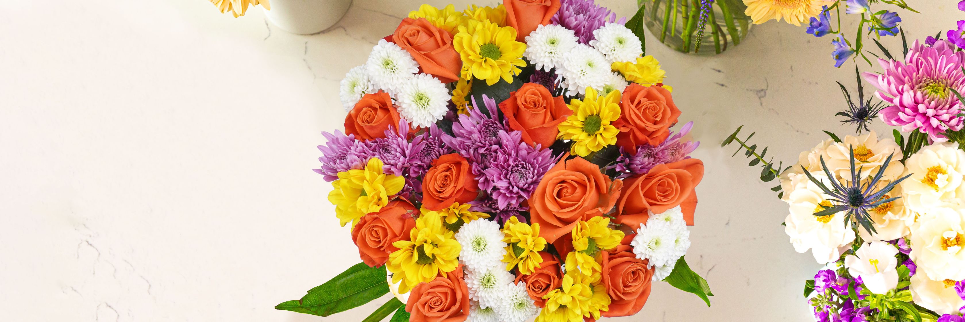 Vibrant floral arrangement featuring orange roses, purple mums, and cheerful daisies.