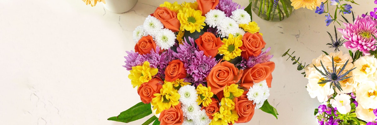 Vibrant floral arrangement featuring orange roses, purple mums, and cheerful daisies.
