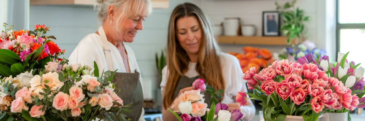 Two florists joyfully arranging vibrant flower bouquets in a bright shop setting.