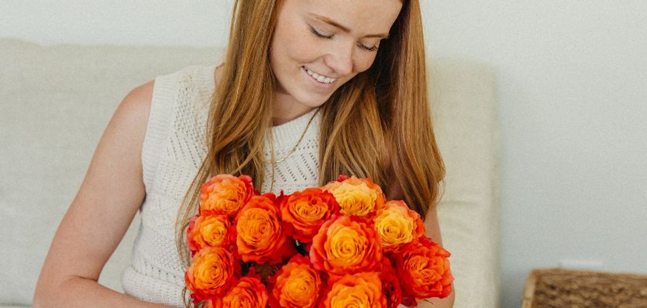 A woman smiles while holding a vibrant bouquet of orange and red roses, radiating joy.