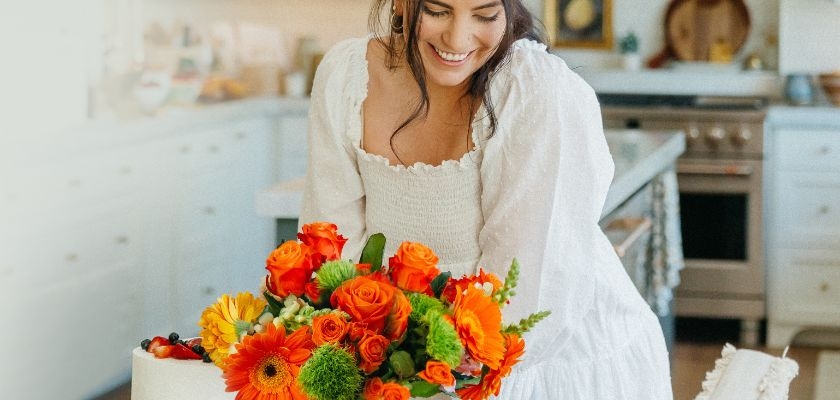 A joyful woman in a white dress arranging a vibrant flower bouquet in a cozy kitchen.
