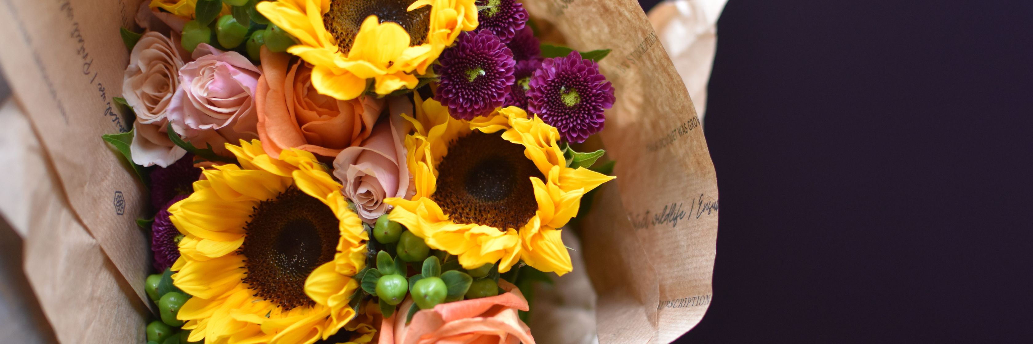 Bright bouquet featuring sunflowers, pink roses, and purple daisies wrapped in elegant paper.