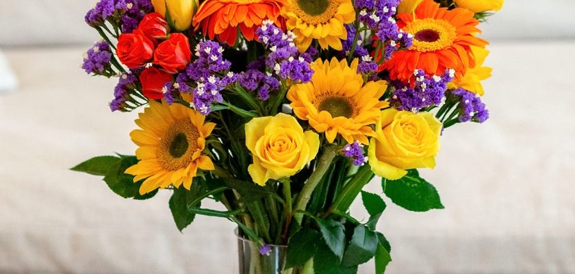 Bright floral bouquet featuring sunflowers, roses, and vibrant gerberas in a glass vase.