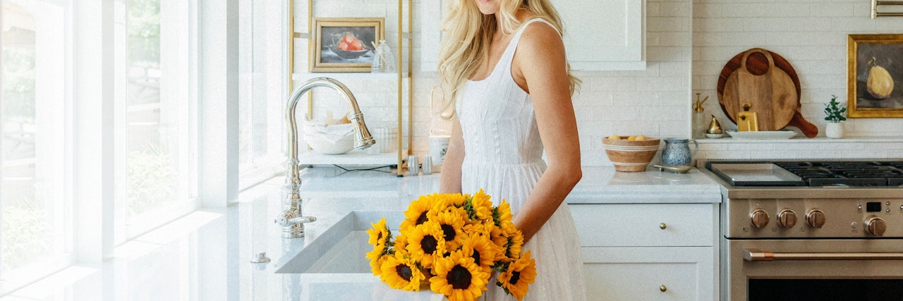 A cheerful woman in a white dress holds a vibrant sunflower bouquet in a bright kitchen.