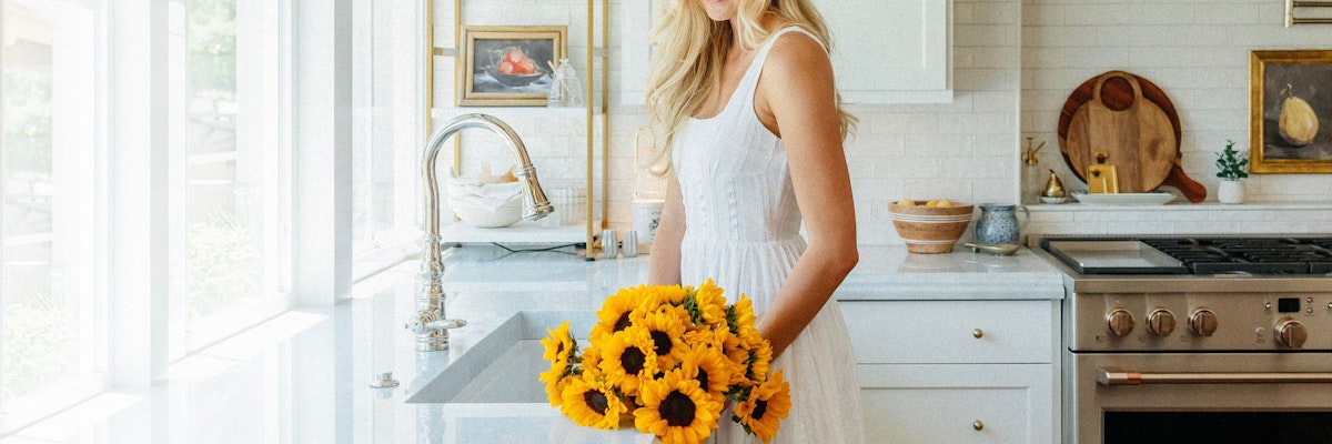 A cheerful woman in a white dress holds a vibrant sunflower bouquet in a bright kitchen.