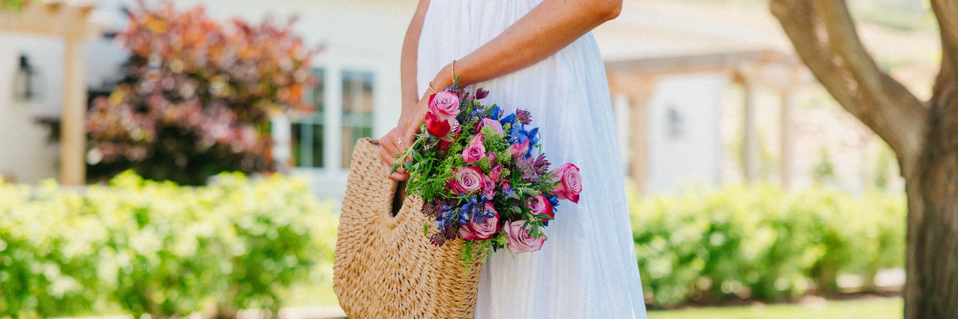 A woman in a white dress carries a vibrant floral bouquet in a woven bag.