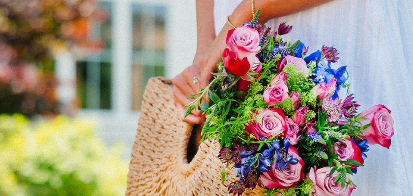 A vibrant bouquet featuring pink roses, colorful blooms, and greenery, held in a woven bag.