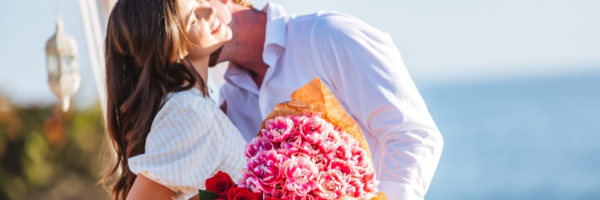 A couple shares a romantic moment by the sea with a vibrant bouquet of pink flowers.