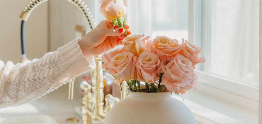 A hand gently adds a pink rose to a lovely bouquet in a classic vase by a sunny window.