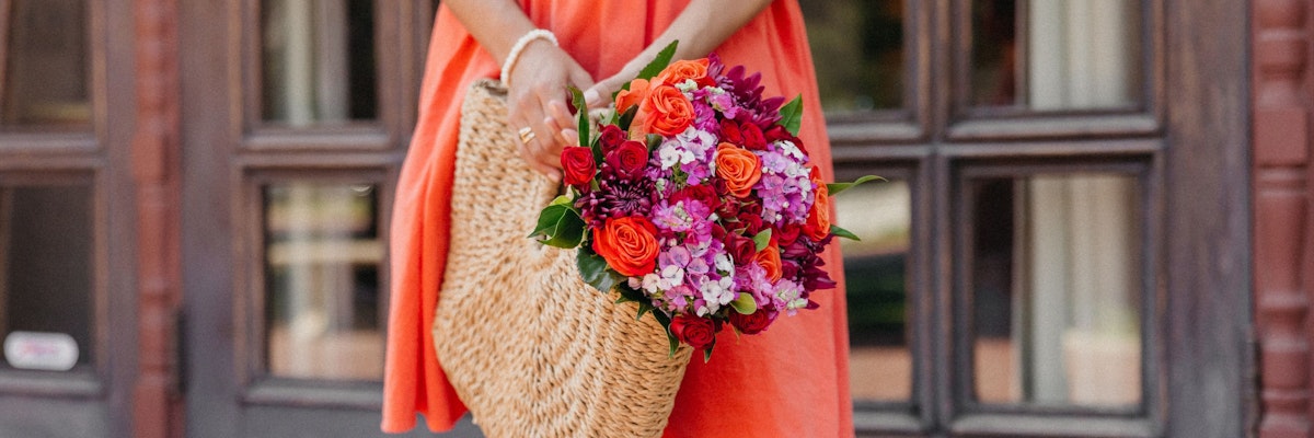 A vibrant floral bouquet of roses and blooms, held in a woven basket against a cheerful backdrop.