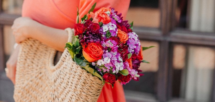 A vibrant flower bouquet featuring red, orange, and purple blooms held in a woven bag.