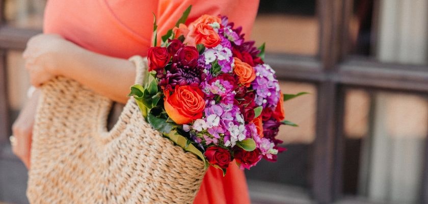 A vibrant flower bouquet featuring red, orange, and purple blooms held in a woven bag.