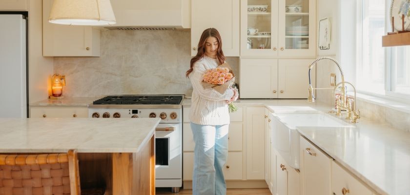 A woman holds a bouquet of orange roses in a bright, airy kitchen setting.