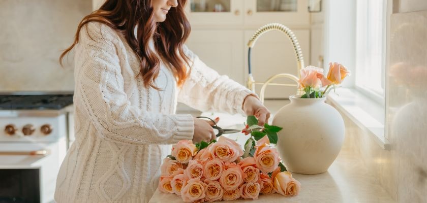 A woman carefully arranges peach roses in a stylish kitchen, creating a vibrant floral display.