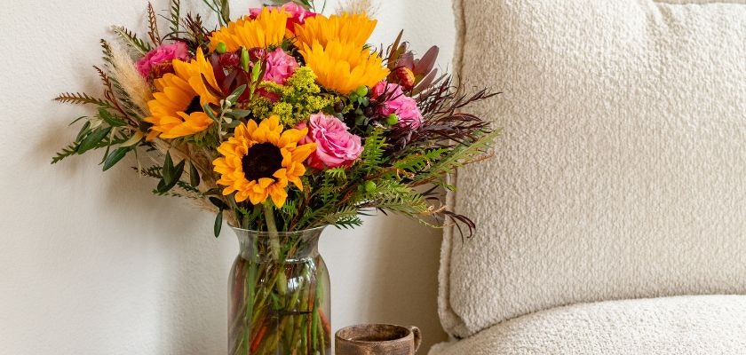 Lively bouquet featuring sunflowers, roses, and greenery in a stylish vase by a cozy setting.
