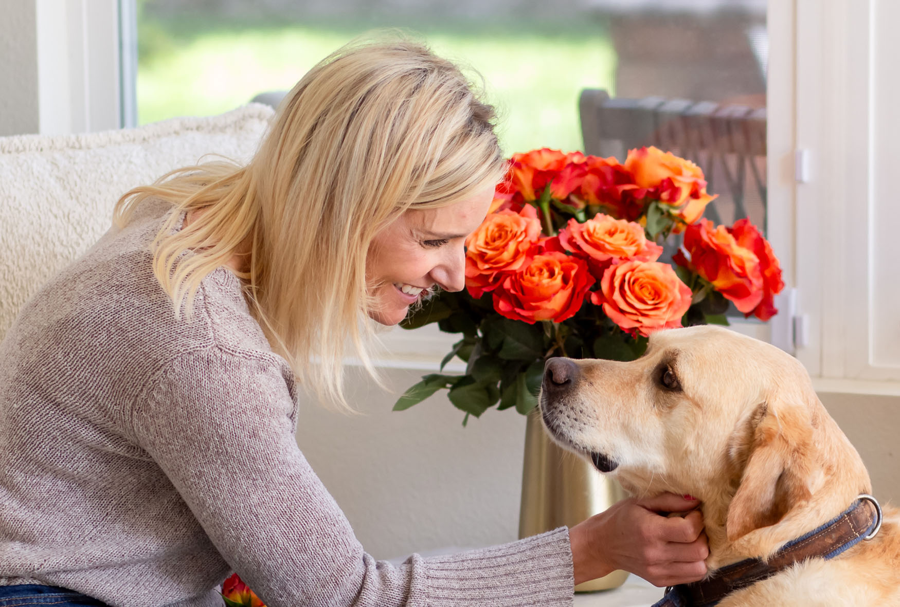 A woman smiles lovingly at her dog beside a vibrant bouquet of orange roses.
