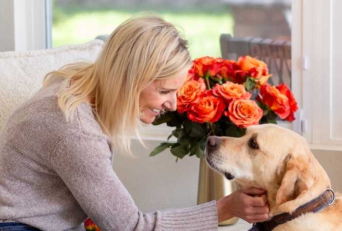 A woman smiles lovingly at her dog beside a vibrant bouquet of orange roses.