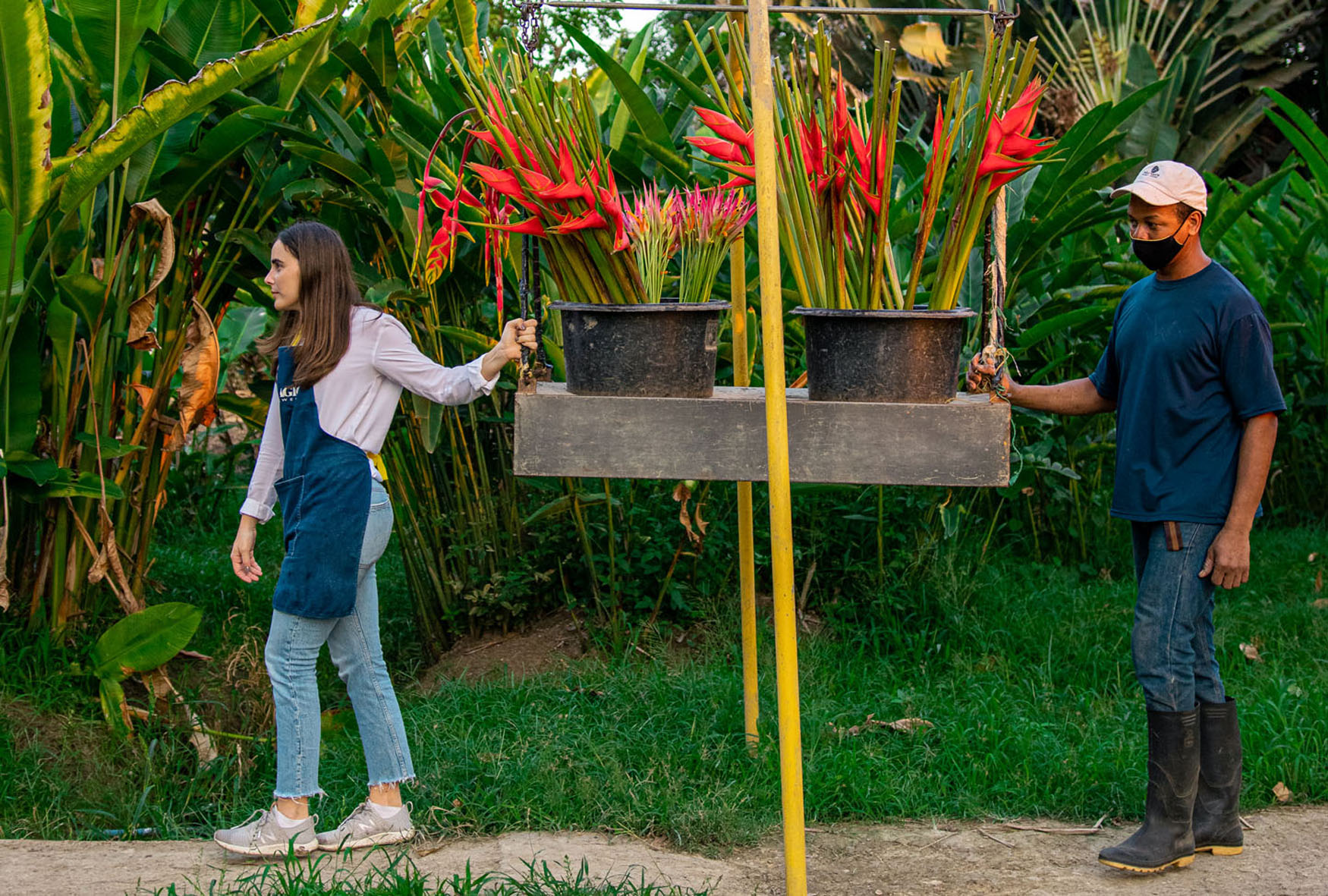 A woman and man carrying vibrant flower pots through a lush green garden path.