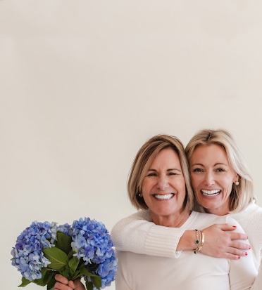 Two smiling women embracing, holding a bouquet of vibrant blue hydrangeas.