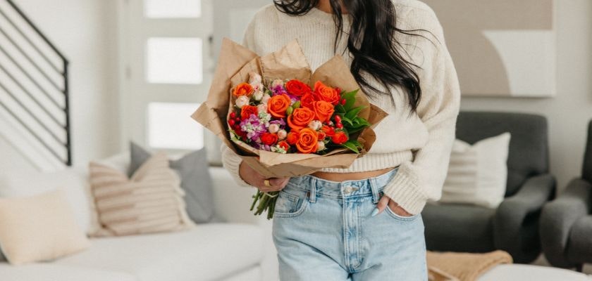A vibrant bouquet of orange roses and assorted flowers, held by a woman in casual attire.
