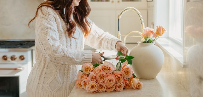 A woman in a cozy sweater arranging soft peach roses in a simple vase at home.