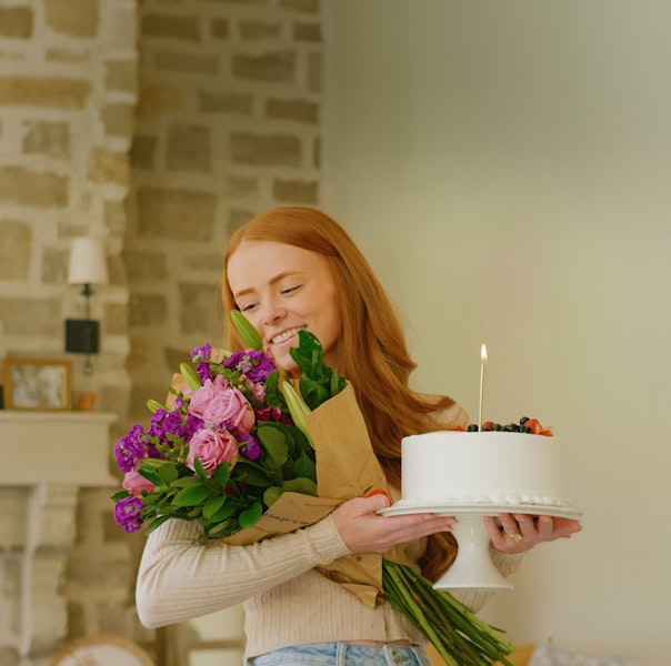A joyful woman holding a beautiful bouquet and a delicious birthday cake.