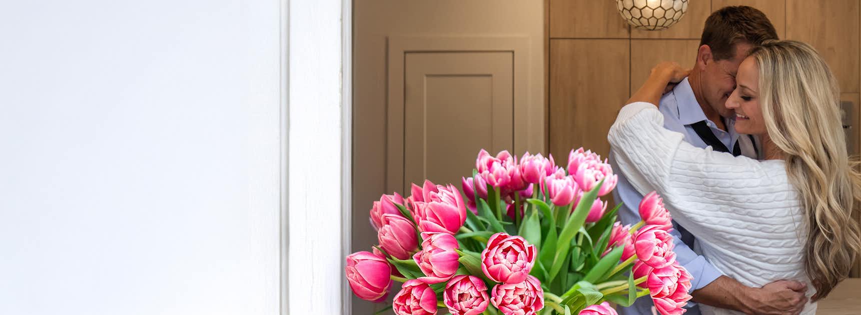 A couple joyfully embracing at home with a vibrant bouquet of pink tulips nearby.