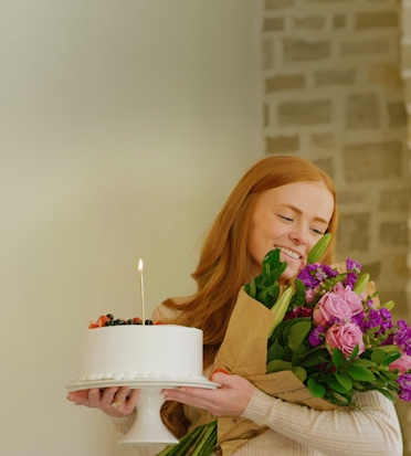 A cheerful redhead holds a cake and a vibrant bouquet, celebrating a special occasion.