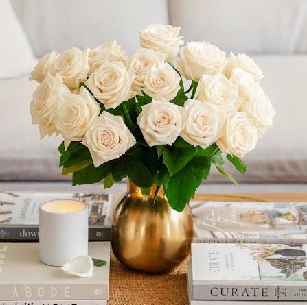Elegant white roses in a gold vase, surrounded by stylish coffee table books and a candle.