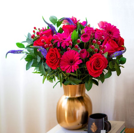 Vibrant bouquet of pink roses, gerbera daisies, and lush greens in a chic golden vase.