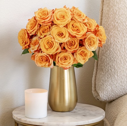 Vibrant orange roses elegantly arranged in a gold vase beside a candle on a marble table.