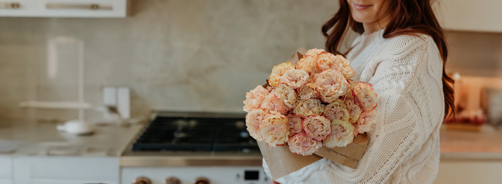 A cozy kitchen scene featuring a woman holding a bouquet of delicate, pastel-colored peonies.