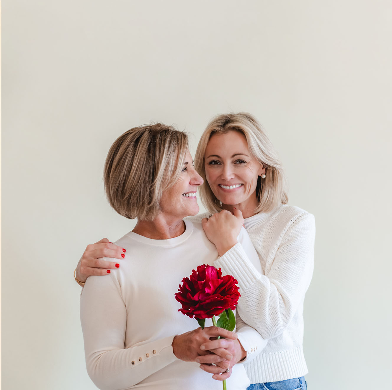 Two smiling women embracing, holding a vibrant red flower against a light background.