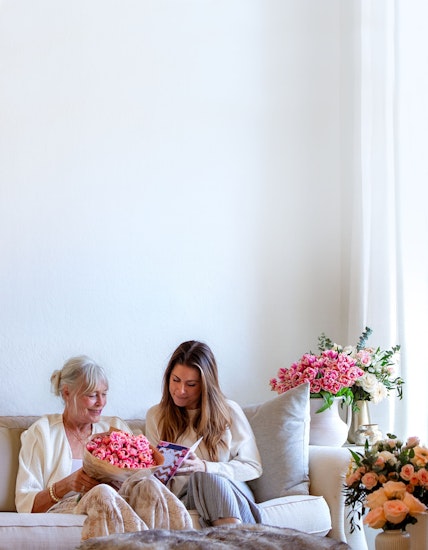 Two women sharing a joyful moment on a couch, surrounded by vibrant flower arrangements.
