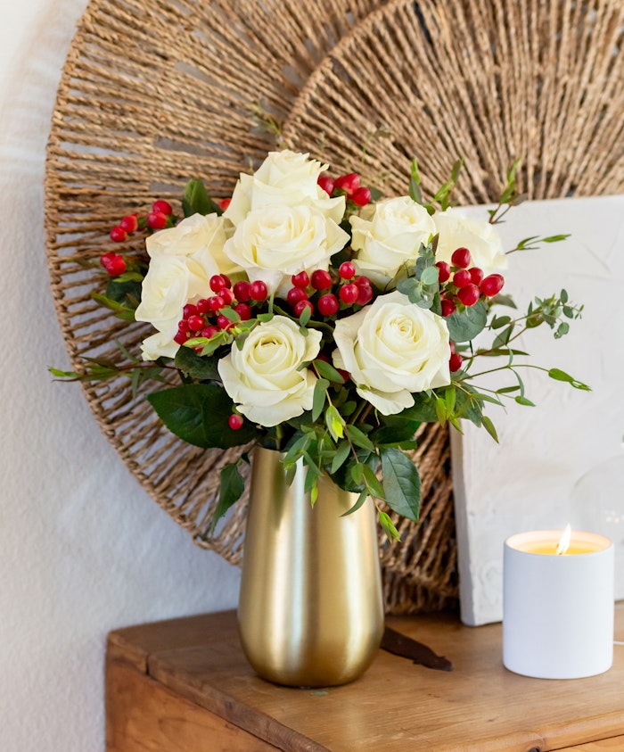 Elegant arrangement of white roses and red berries in a chic gold vase, perfect for decor.