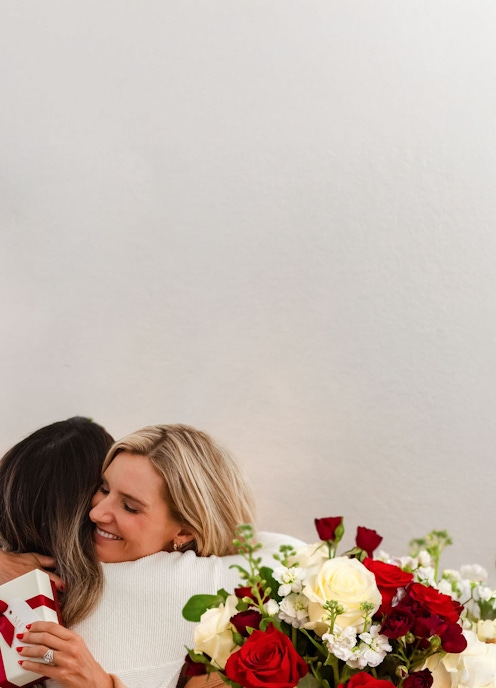 Two women share a warm embrace, surrounded by a vibrant bouquet of red and white roses.