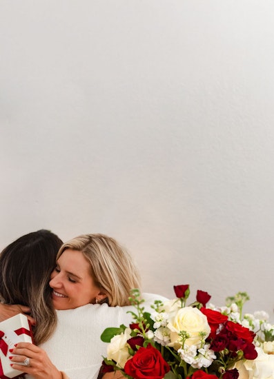 Two women share a warm embrace, surrounded by a vibrant bouquet of red and white roses.
