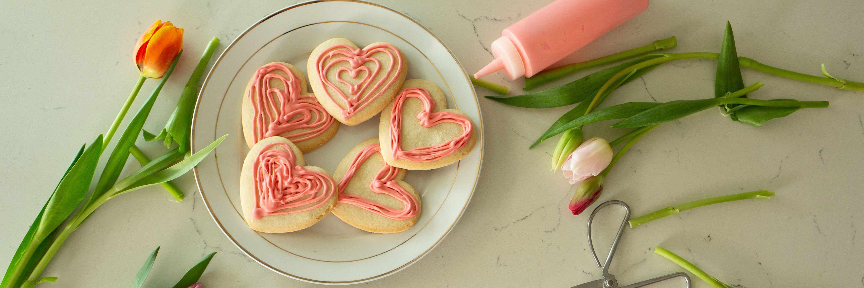 Decorative heart-shaped cookies with pink icing surrounded by fresh tulips on a marble table.
