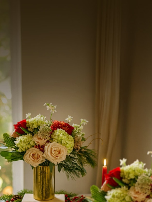 Elegant floral arrangement featuring roses, hydrangeas, and festive greenery in a gold vase.