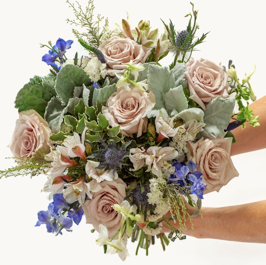 Elegant bouquet featuring soft pink roses, blue delphiniums, and lush greenery, beautifully arranged.
