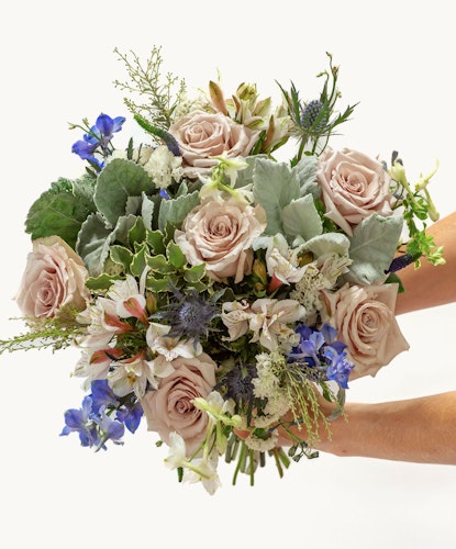 Elegant bouquet featuring soft pink roses, blue delphiniums, and lush greenery, beautifully arranged.